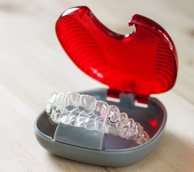 Open orthodontic retainer case with clear retainer inside, red lid raised, gray base visible.